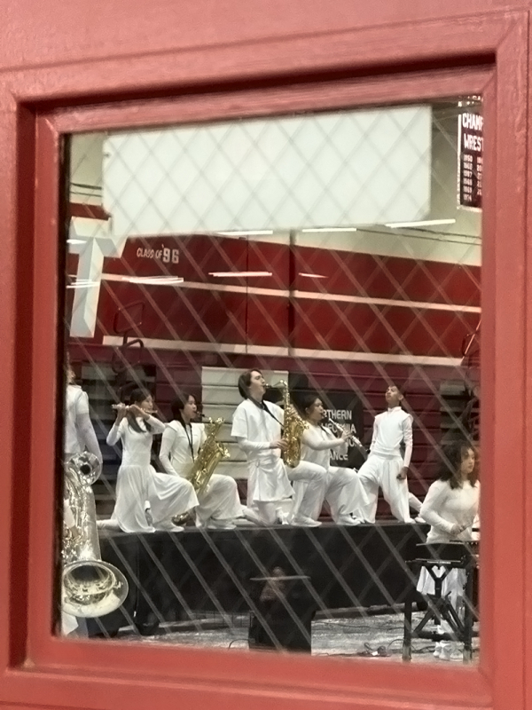 Various woodwind players kneeling on a raised stage viewed through the window in a door.