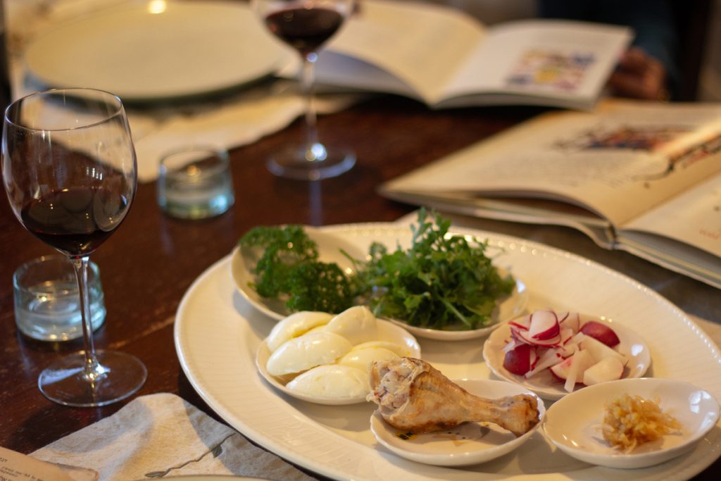 A Seder plate with various foods by a wine glass and a Passover book.