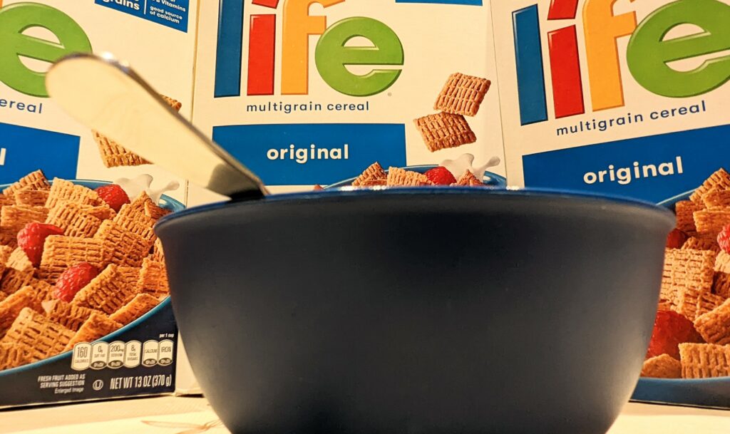A bowl with a spoon with a wall of Life cereal boxes behind it.