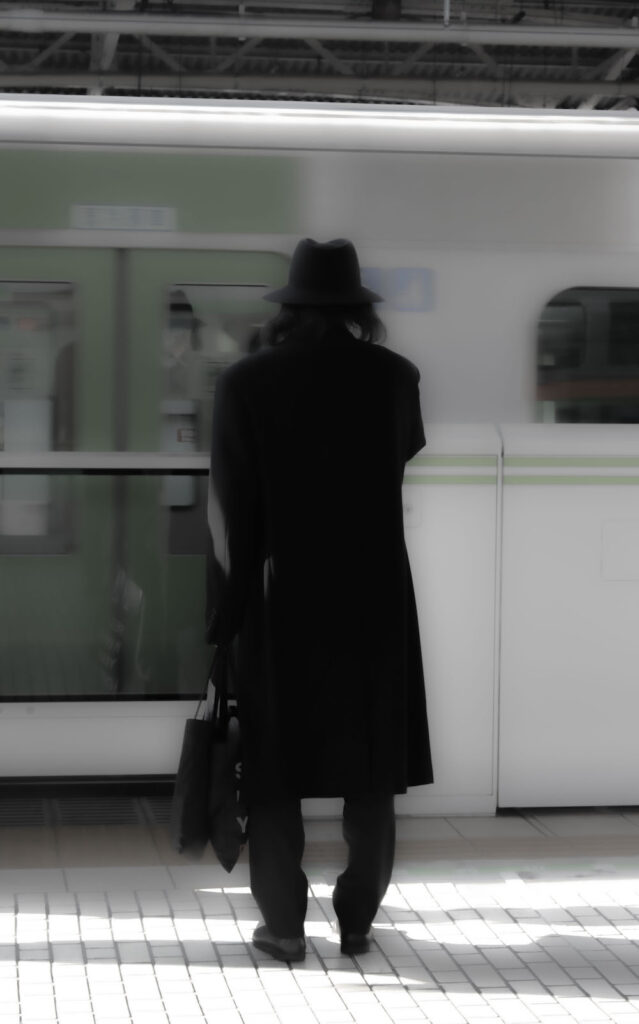 a photo of a man standing on a train platform as a train goes by, wearing a long coat and a hat. 