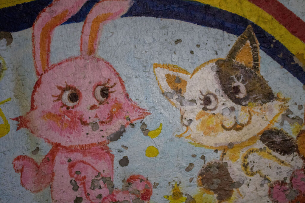 a photo of an old chipped mural of a happy cartoon bunny and cat in a flower field with a rainbow above them. 