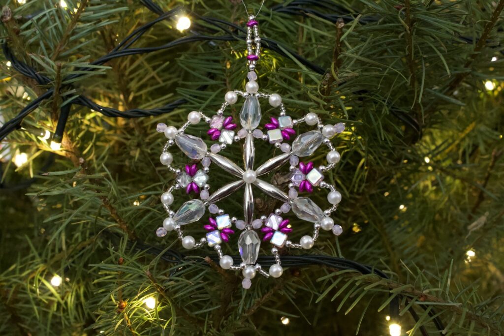 Snowflake shaped ornament made of beads hung in the branches of a Christmas tree with lights
