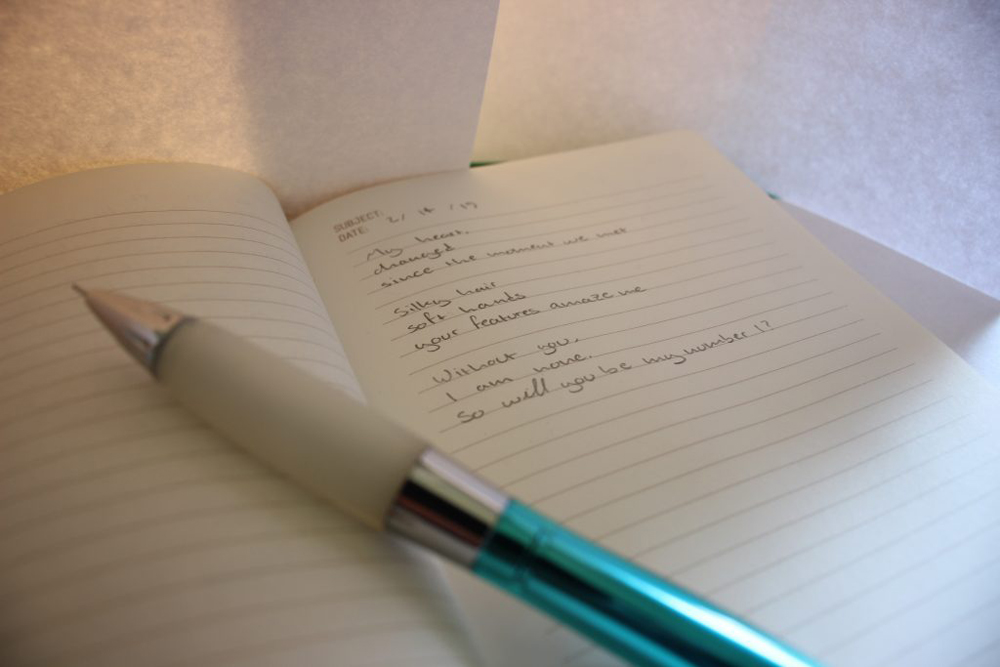 The image pictures an opened notebook with a poem written in it and a pencil laid out over the notebook. 