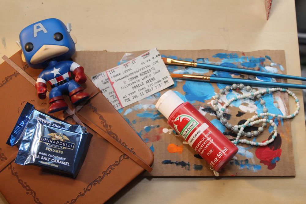Photo shows a Captain America Pop figurine, Ghirardelli chocolate squares, a sketchbook, a concert ticket, a paint bottle, paint brushes, a piece of cardboard with all colors of paint allover it and a blue and white beaded necklace and bracelet. 