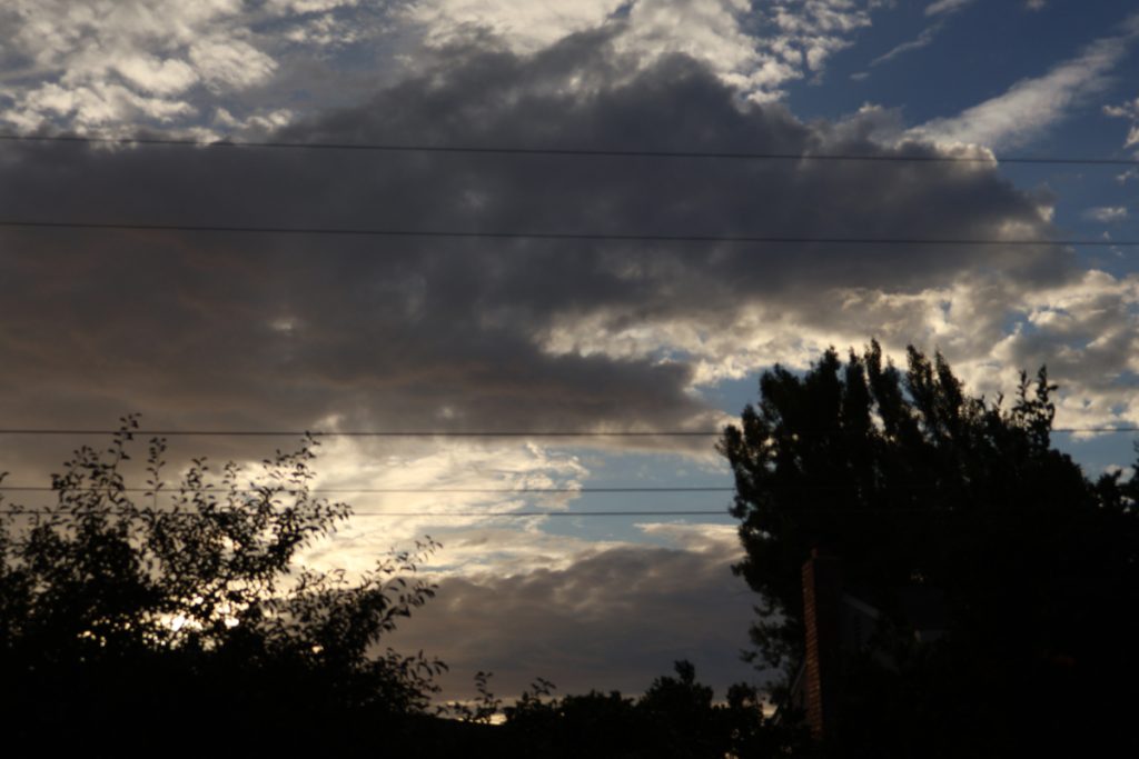 image of two darkened trees in foreground, small powerlines cross the entire image. Large clouds billow across the backround with the sky visible more on the right side. Light from the sun peeks through the bottom left tree