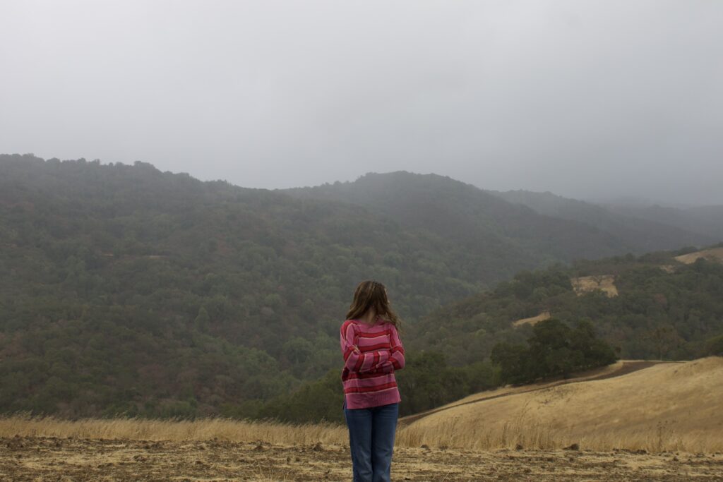 I (Sheer) am standing center frame looking off to the side. Behind me are large hills covered by fog and rain.