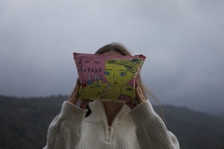 A vibrant pencil case that says, "I am an Artist", is covering my face. The background are foggy hills.