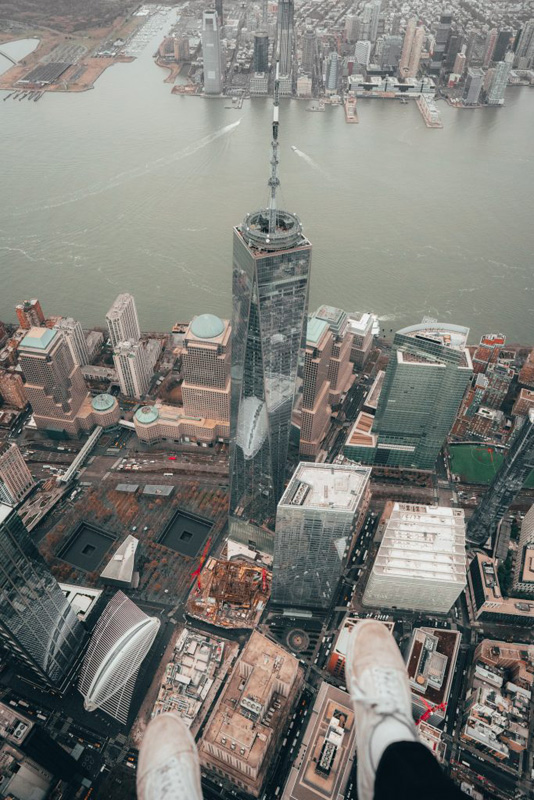  An aerial photo looking down at the One World Trade Center in New York City. With the river in the background. Feet are hanging from the helicopter.