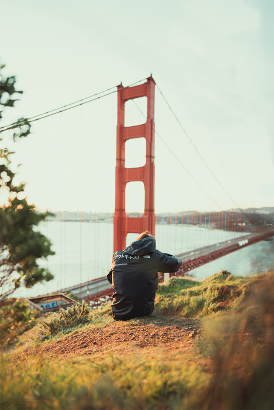 the photo shows a teen taking a photo of the golden gate bridge in San Francisco, the foreground being grass and leaves.