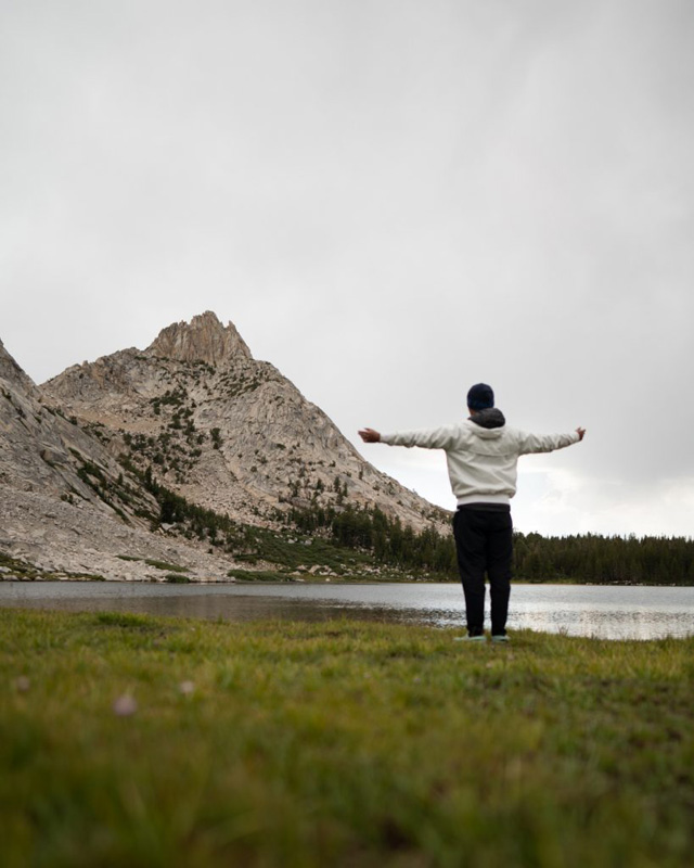 the photo shows a young man with his hands up on this sides looking out at a view with a peak of a mountain and a small lake right in front of him.