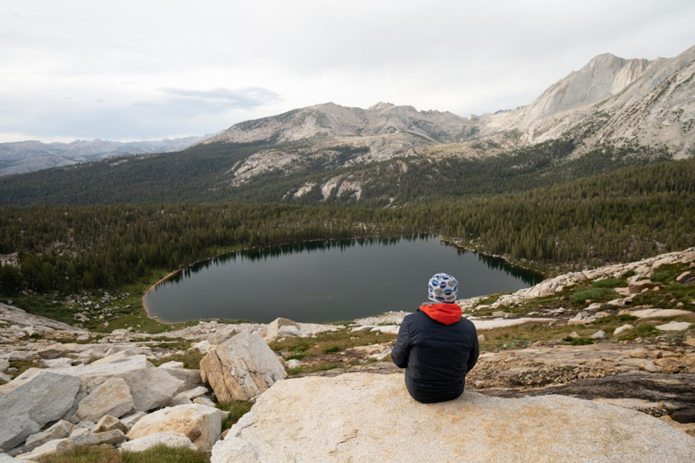 The photo shows a man sitting on a rock in the foreground, while in the background he's looking out into a lake and a bunch of mountain peeks.