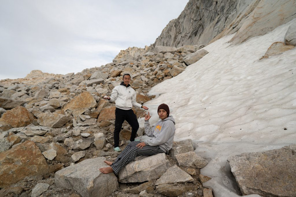 the photo shows 2 teenagers standing and sitting next to a pack of ice, surrounded by a lot of big rocks and boulders.