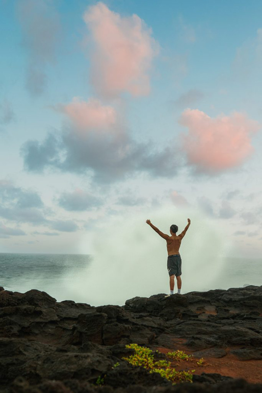 A picture of a man standing near the water with his hands up in the sky. There is water splashing in front of him.