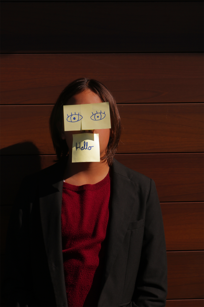 In the image there is a kid with 3 post-its on his face. 2 on top of his eyes with eyes drawn on them, and finally one on top of his mouth with the word hello written on it. 
