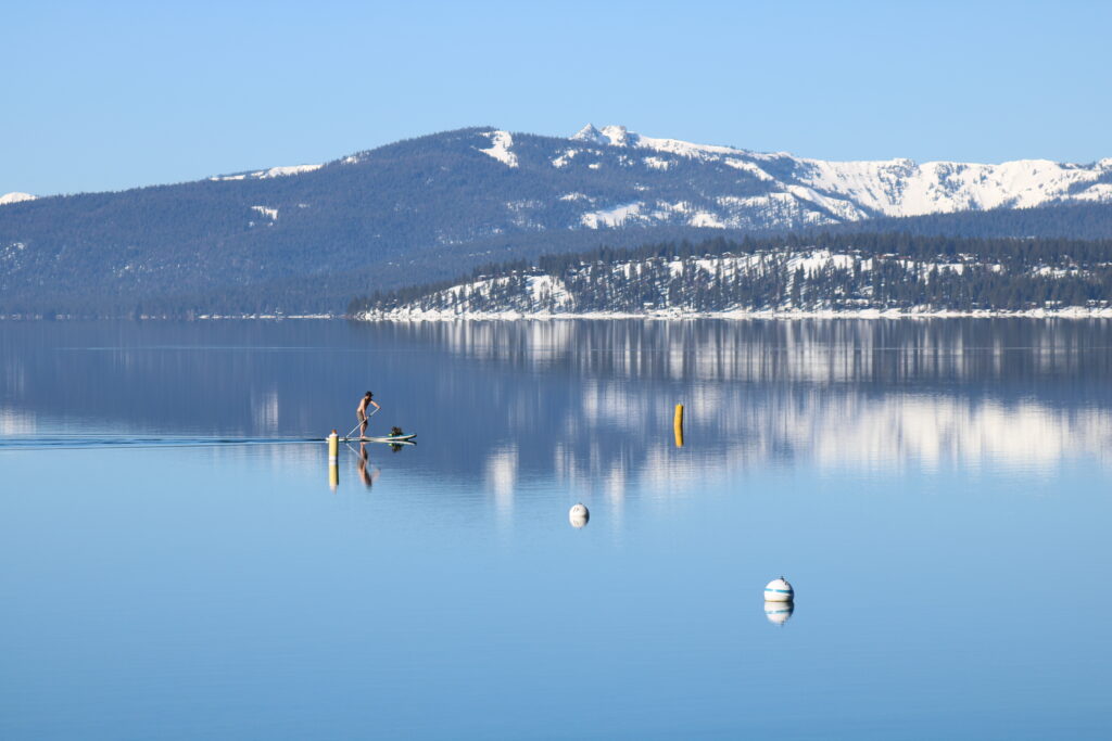 Background: snow covered mountains that reach the calm lake

There is a person that is on a stand up paddle board, the leave ripples in the water behind them