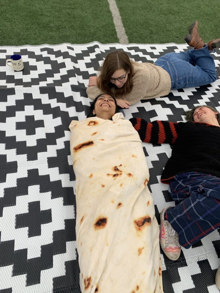 3 people laughing on the ground. One is wrapped in a burrito blanket in the middle. 
